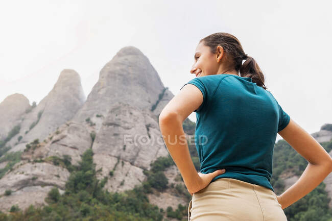 Von unten Rückansicht einer fröhlichen Reisenden mit den Händen auf den Hüften, die Montserrat mit Bäumen betrachtet, während sie bei einem Ausflug in Spanien wegschaut — Stockfoto