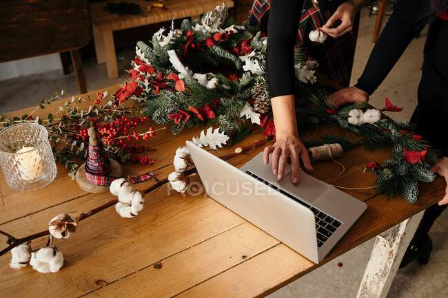 Обрезали неузнаваемых женщин-флористок, стоящих за столом и использующих ноутбук, делая рождественские украшения в стильной цветочной студии — стоковое фото