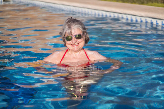 Alegre anciana hembra con pelo gris nadando en la piscina y sonriendo brillantemente a la cámara con gafas de sol - foto de stock