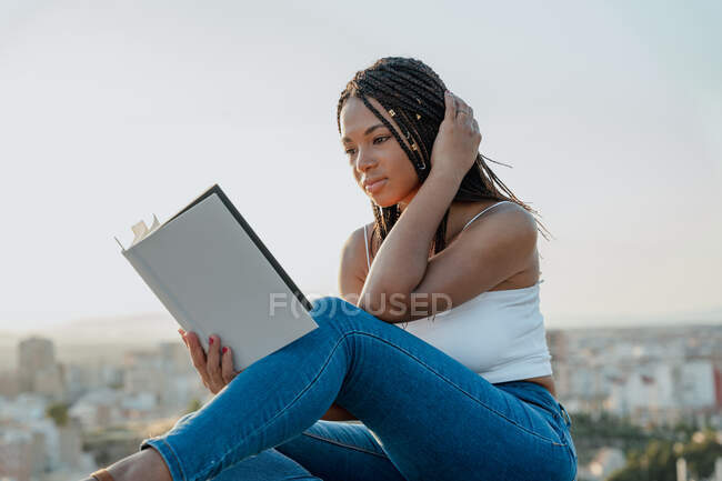 Mujer étnica de moda en sandalias leyendo libro de texto mientras está sentada con las piernas cruzadas en la valla en la ciudad - foto de stock