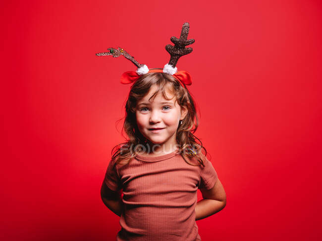 Alegre niña en ropa casual y diadema de ciervo festivo mirando a la cámara durante la celebración de Navidad contra el fondo rojo - foto de stock