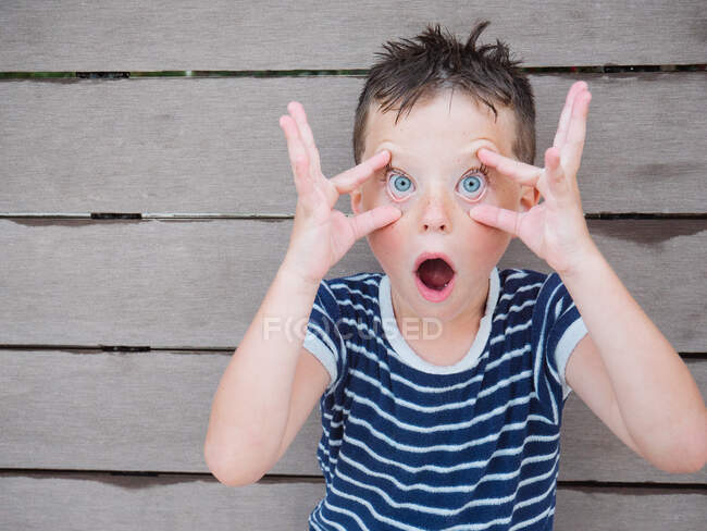 Brincalhão garoto surpreso com cabelos molhados abrindo os olhos com as mãos enquanto expressa espanto e olhando para a câmera — Fotografia de Stock