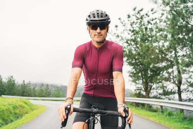 Deportista adulto en gafas de sol de ciclismo y casco sentado en bicicleta de carretera en la carretera rural a la luz del día - foto de stock