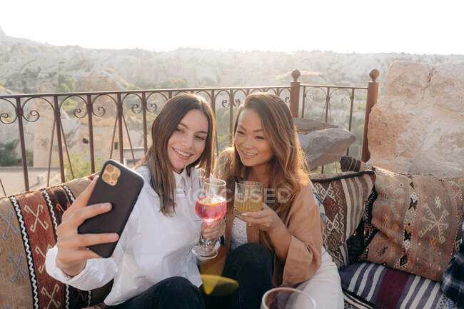 Angle élevé d'amies heureuses ethniques assises sur des oreillers tout en prenant un autoportrait sur un téléphone portable près de la table avec appareil photo instantané et cocktail en terrasse en Cappadoce, Turquie — Photo de stock