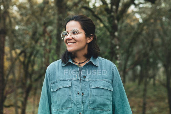 Positive ethnic female in eyeglasses wearing denim shirt standing among trees and looking away with toothy smile — Stock Photo
