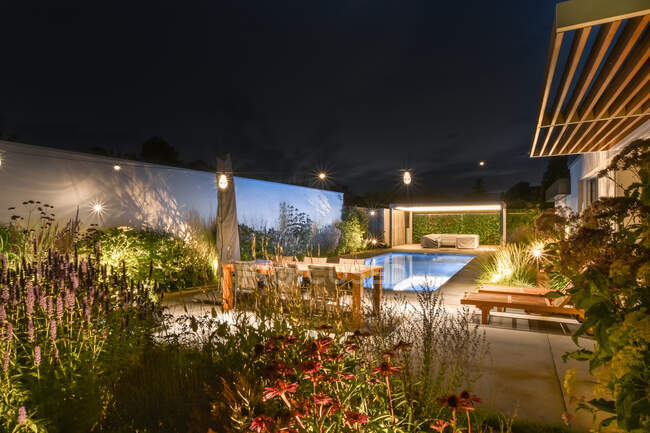Amplio patio trasero de villa contemporánea con mesa de madera y tumbonas y plantas verdes colocadas cerca de la piscina contra el cielo nocturno sin nubes - foto de stock