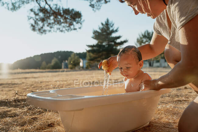 Récolté maman méconnaissable baignade tout-petit enfant tout en étant assis sur la prairie sous un ciel brillant le jour ensoleillé — Photo de stock