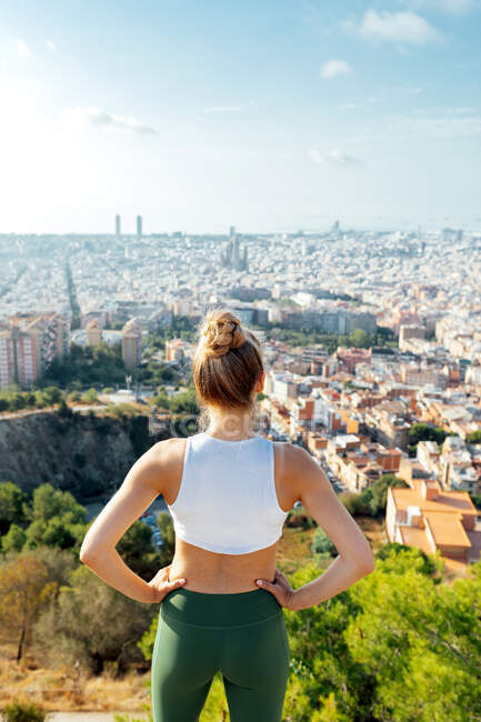 Vista posterior de una atleta anónima con las manos en la cintura admirando la ciudad de verano a la luz del sol - foto de stock