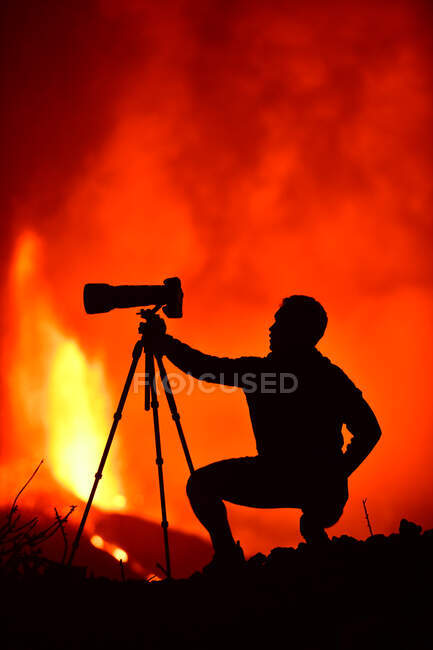 Seitenansicht der Silhouette eines Mannes, der sich hinduckt und mit Teleobjektiv und Stativ die Lavaexplosion auf den Kanarischen Inseln La Palma 2021 fotografiert — Stockfoto