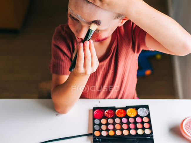 Charming child with makeup applicator touching head while looking away at table with eyeshadow palette — Stock Photo