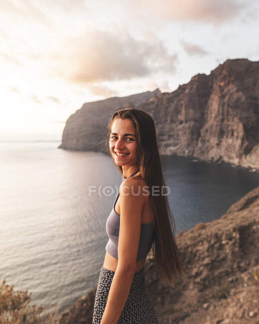 Vista lateral da fêmea sorridente com cabelos longos no cume olhando para a câmera contra o oceano ao pôr-do-sol em Tenerife Espanha — Fotografia de Stock