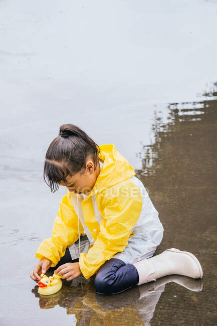 Charmant enfant ethnique en imperméable jouant avec des canards en plastique se reflétant dans la flaque ondulée par temps pluvieux — Photo de stock