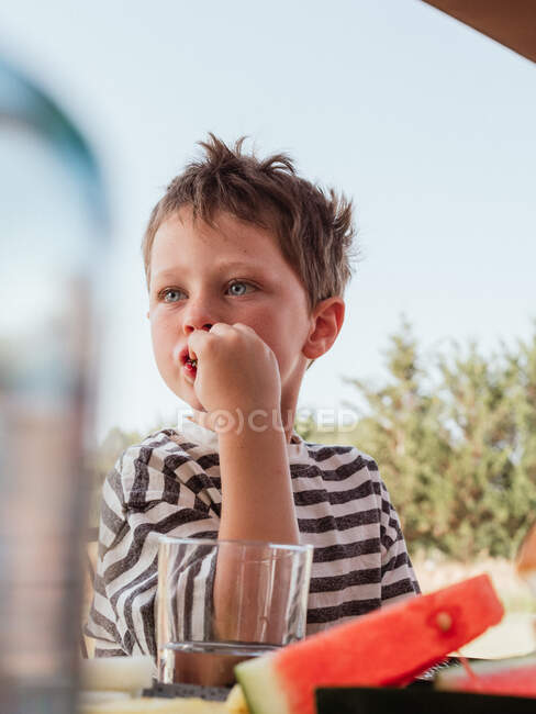 Baixo ângulo de uma criança pequena e fofa sentada à mesa no pátio no verão e comendo melancia doce fresca enquanto olha para longe — Fotografia de Stock