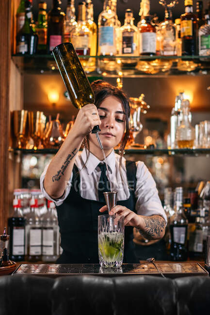 Joven camarera sirviendo bebida alcohólica de botella en jigger mientras prepara cóctel en el mostrador en el bar - foto de stock