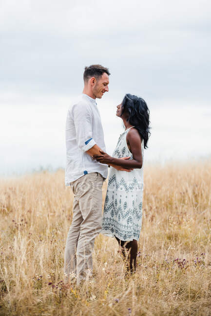 Vista lateral do homem sorridente abraçando a namorada indiana enquanto olham um para o outro no campo sob o céu nublado — Fotografia de Stock