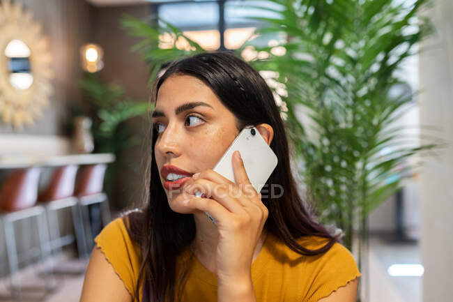 Mujer freelancer seria sentada en la mesa de madera en la cafetería y teniendo una llamada telefónica mirando hacia otro lado - foto de stock
