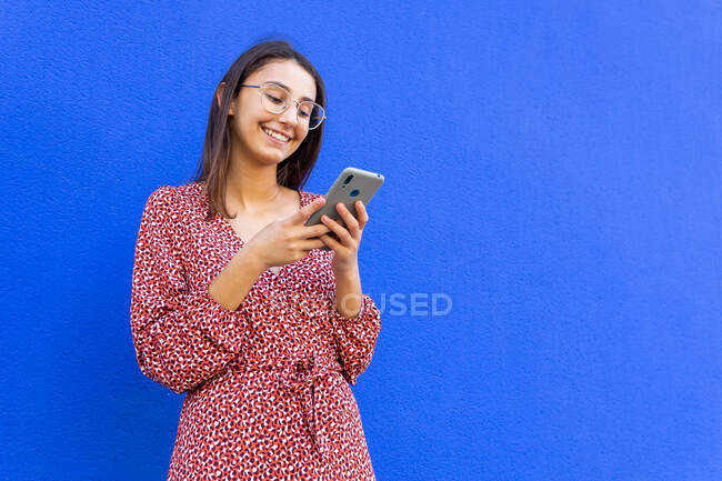 Улыбающаяся женщина в платье и очках стоит у синей стены и днем пользуется смартфоном — стоковое фото