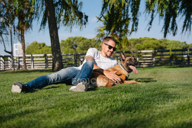 Jovem barbudo macho em óculos de sol deitado no prado contra cão de raça pura no parque no dia de verão — Fotografia de Stock