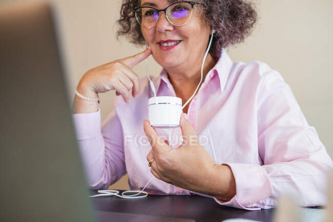 Crop smiling senior female entrepreneur in eyeglasses and earphones with jar of cream touching cheek during video chat on netbook — Stock Photo