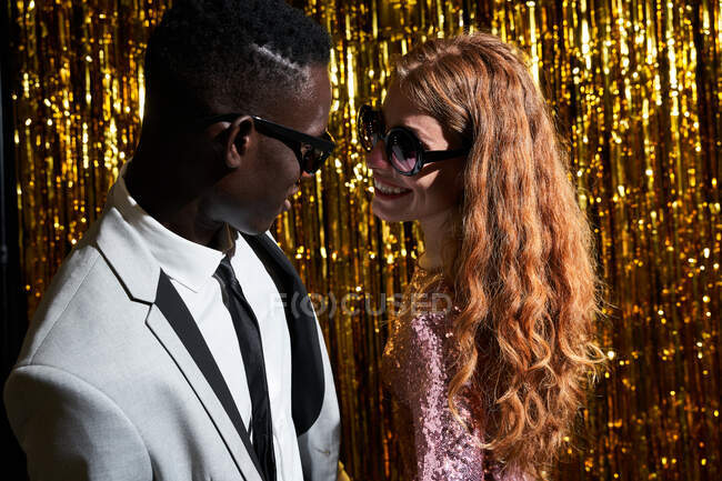 Contenido joven pareja multiétnica en gafas de sol modernas mirándose en el momento del beso contra el oropel durante la celebración de Nochevieja - foto de stock
