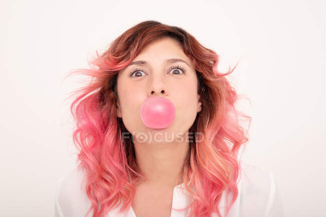 Femme gaie avec des cheveux roses soufflant gomme à bulles et regardant la caméra sur fond clair — Photo de stock