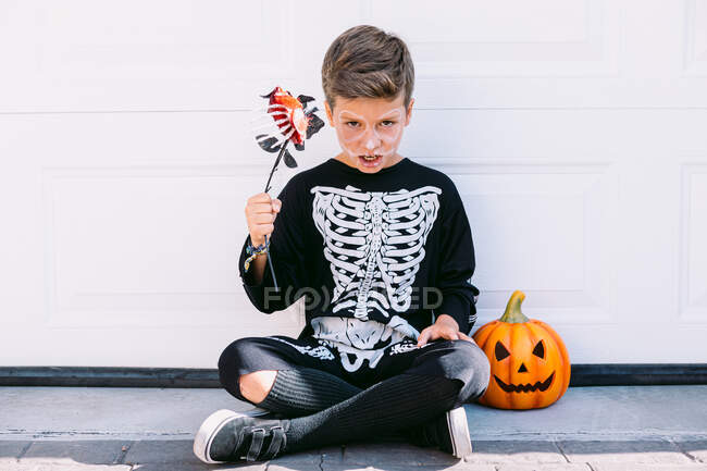 Cuerpo completo de niño preadolescente espeluznante en traje de esqueleto negro con maquillaje sosteniendo la flor y mirando a la cámara mientras está sentado cerca de la linterna de calabaza contra la pared blanca - foto de stock