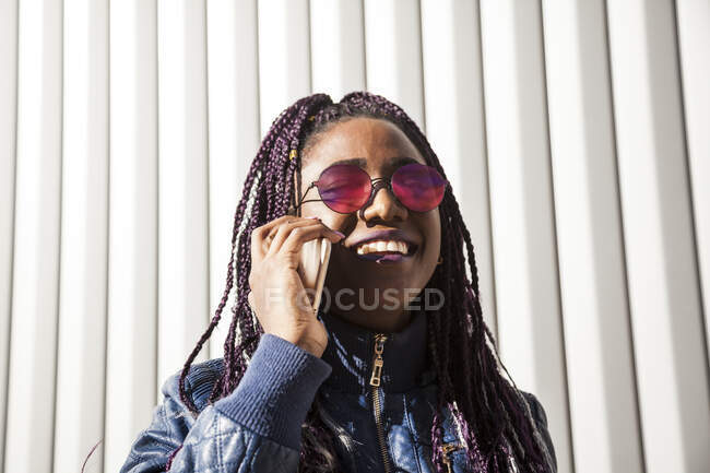 Joven mujer afroamericana alegre y elegante con trenzas afro con chaqueta de moda y gafas de sol hablando por teléfono móvil mientras está de pie cerca de la pared del edificio urbano - foto de stock