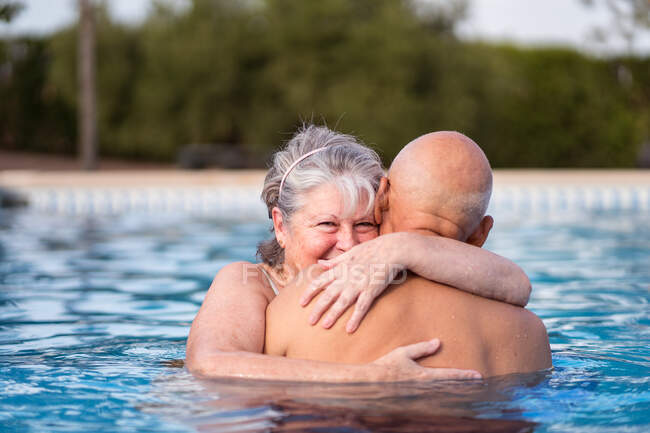 Sonriente mujer de pelo gris abrazando al hombre calvo sin camisa mientras nadan juntos en agua limpia de la piscina - foto de stock