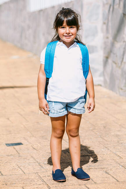 Школярка з рюкзаком на тротуарі дивиться на камеру на сонячному світлі — стокове фото