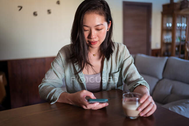 Jovem fêmea étnica com vidro de café surf internet no celular à mesa na sala da casa — Fotografia de Stock