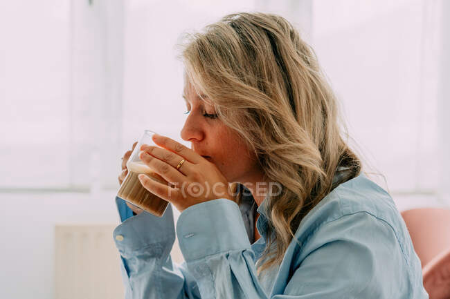 Side view of reflective adult female with wavy hair drinking tasty coffee in house — Stock Photo