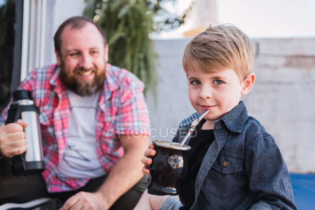 Enfant sincère avec calebasse gourde de boisson infusée contre papa barbu joyeux avec thermos sur fond flou — Photo de stock