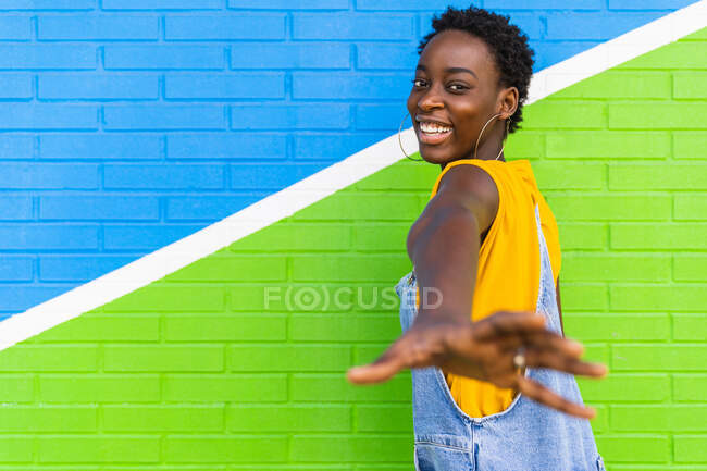 Visão traseira da fêmea afro-americana encantada ouvir parede colorida e chegar a mão para câmera — Fotografia de Stock