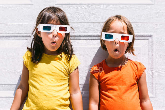 Bonito meninas surpreendidas em roupas coloridas casuais e óculos tridimensionais em pé no fundo da parede branca — Fotografia de Stock