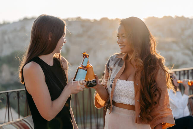Vista lateral de jóvenes amigas multiétnicas positivas sonriendo brillantemente y tintineando botellas de cerveza mientras disfrutan de un agradable tiempo juntos al atardecer en el bar terraza en Capadocia, Turquía - foto de stock