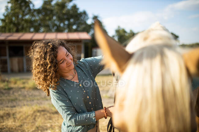 Contenido maduro hembra acariciando semental hocico con abrigo marrón liso contra el heno en la escuela de equitación en día soleado - foto de stock