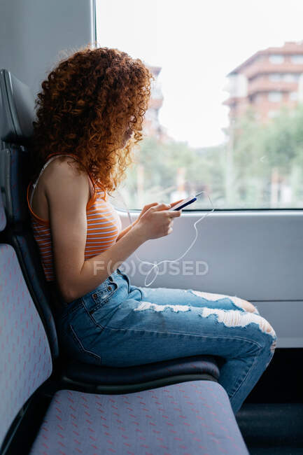 Заинтересованная женщина с вьющимися волосами в разорванных джинсах, отправляющая смс по мобильному телефону во время поездки на поезде днем — стоковое фото