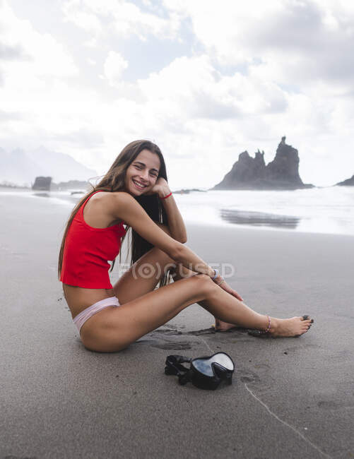 Side view of cheerful female teenager in bikini resting on ocean coast while looking at camera against mounts in Tenerife — Stock Photo