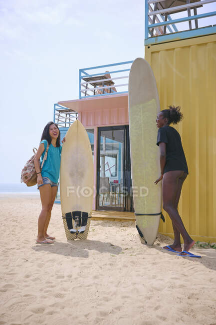 Zufriedene junge multirassische Sportlerinnen mit Longboard und Surfbrett stehen am Sandstrand gegen Baumaßnahmen unter bewölktem Himmel — Stockfoto