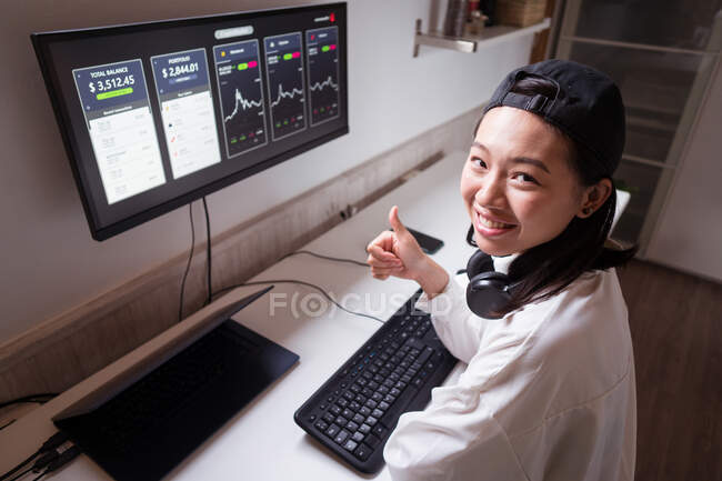 Зверху усміхнена етнічна жінка-брокер друкує на клавіатурі проти монітора з графікою і показує, як жест, дивлячись на камеру вдома — стокове фото