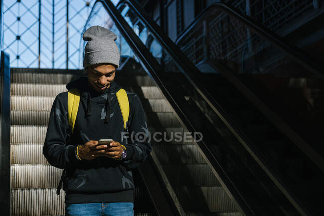 Side view of delighted African American male hipster with backpack standing on moving stairs surfing mobile phone — Stock Photo