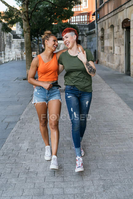 Vista posterior de mujeres homosexuales jóvenes con tatuajes abrazándose mientras caminan por la pasarela en la ciudad - foto de stock