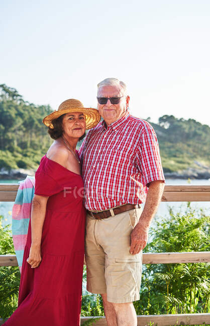 Coppia anziana in piedi uno accanto all'altro guardando la fotocamera e godendo della vista sul mare nella giornata di sole — Foto stock