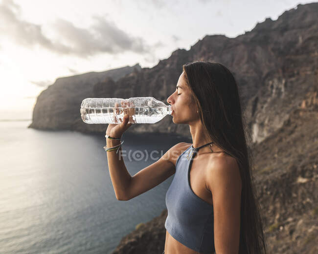 Vue latérale d'une adolescente consciente aux yeux fermés et aux cheveux longs buvant de l'eau depuis une bouteille sur le mont contre l'océan à Ténérife — Photo de stock