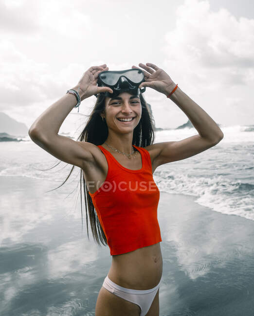 Cheerful female teenager in bikini and crop top with diving mask looking at camera against stormy sea in Tenerife Spain — Stock Photo