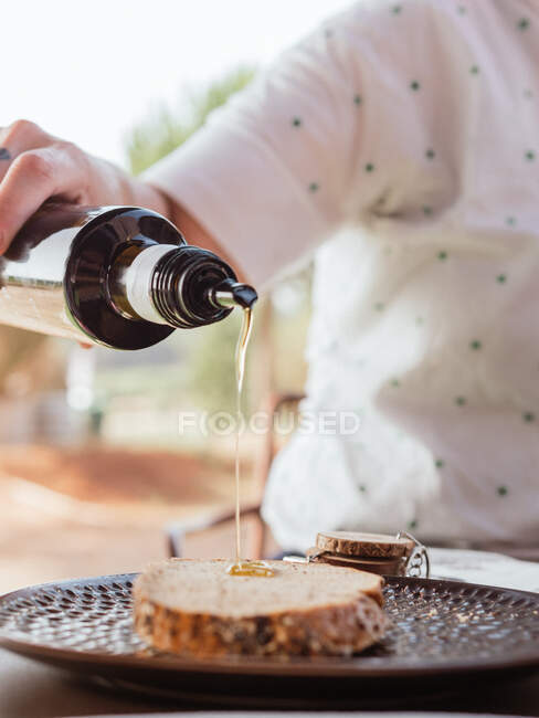Cultivo irreconocible persona añadiendo jarabe dulce en rebanada de pan en el plato colocado en la mesa para el desayuno en la terraza de verano - foto de stock