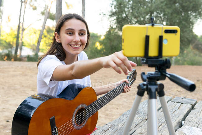 Sorrindo feminino adolescente guitarrista gravação de vídeo no celular moderno no tripé no parque no fundo borrado — Fotografia de Stock