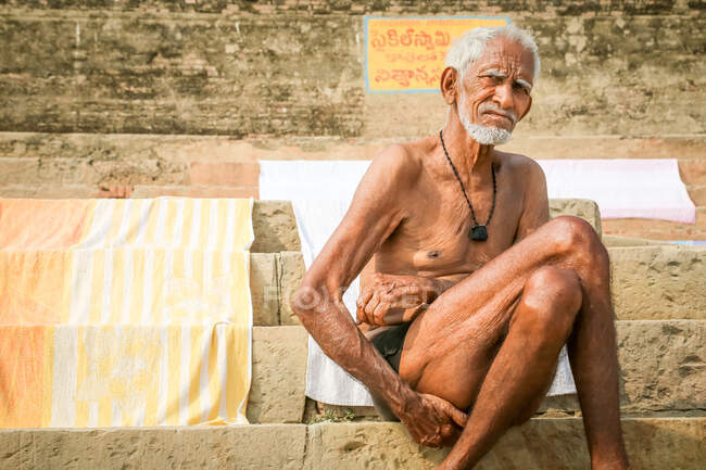 INDIA, VARANASI - 27 DE NOVIEMBRE DE 2015: Hombre indio sin camisa con el pelo gris sentado en escaleras de piedra y mirando a la cámara - foto de stock