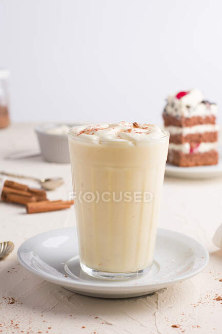 Glass of milk punch with cinnamon powder on whipped egg white against cake piece on cafeteria table on light background — Stock Photo