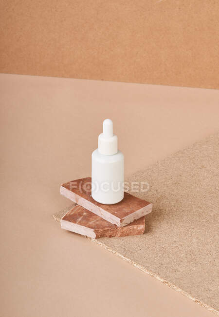 Pequeña botella blanca de suero cosmético colocada en piezas de piedra de mármol marrón apilado sobre tela sobre fondo beige - foto de stock